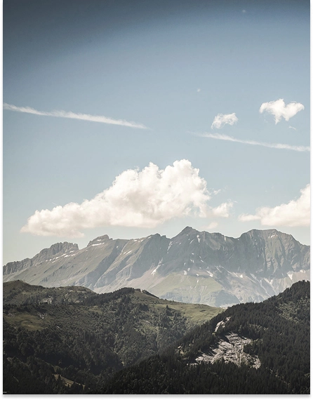Panorama de sommets alpins avec des traces de neige résiduelle, entourés de forêts de sapins et de pâturages verts, sous un ciel bleu avec quelques nuages. Paysage estival des Alpes françaises.