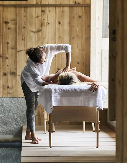 Masseuse en blouse blanche prodiguant un massage du dos à une cliente allongée sur une table en bois drapée d'une serviette blanche. Cabine de soin en bois de mélèze avec lumière naturelle latérale, soubassement en pierre visible. Ambiance spa alpin serein et épuré.