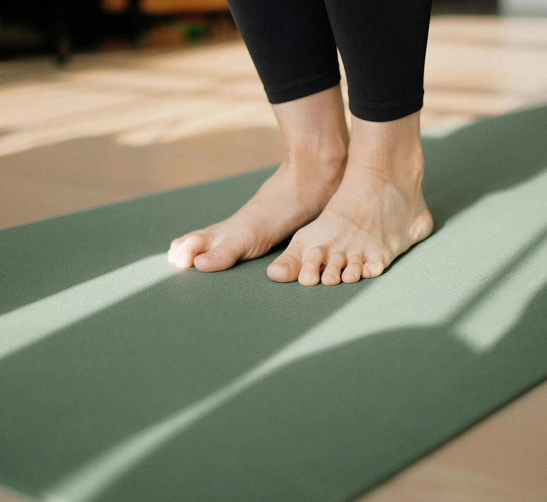Gros plan sur les pieds nus d'une personne en legging noir debout sur un tapis de yoga vert, traversé par des jeux d'ombres et de lumière naturelle. Pratique du yoga ou du fitness en intérieur.