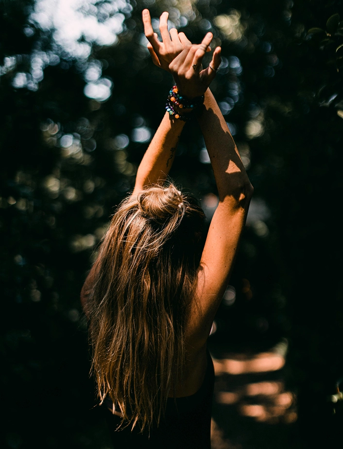 Vue de dos d'une femme aux longs cheveux blonds levant les bras vers le ciel, bracelets de perles colorées aux poignets, entourée d'un sous-bois en lumière tamisée. Atmosphère méditative et connexion avec la nature.
