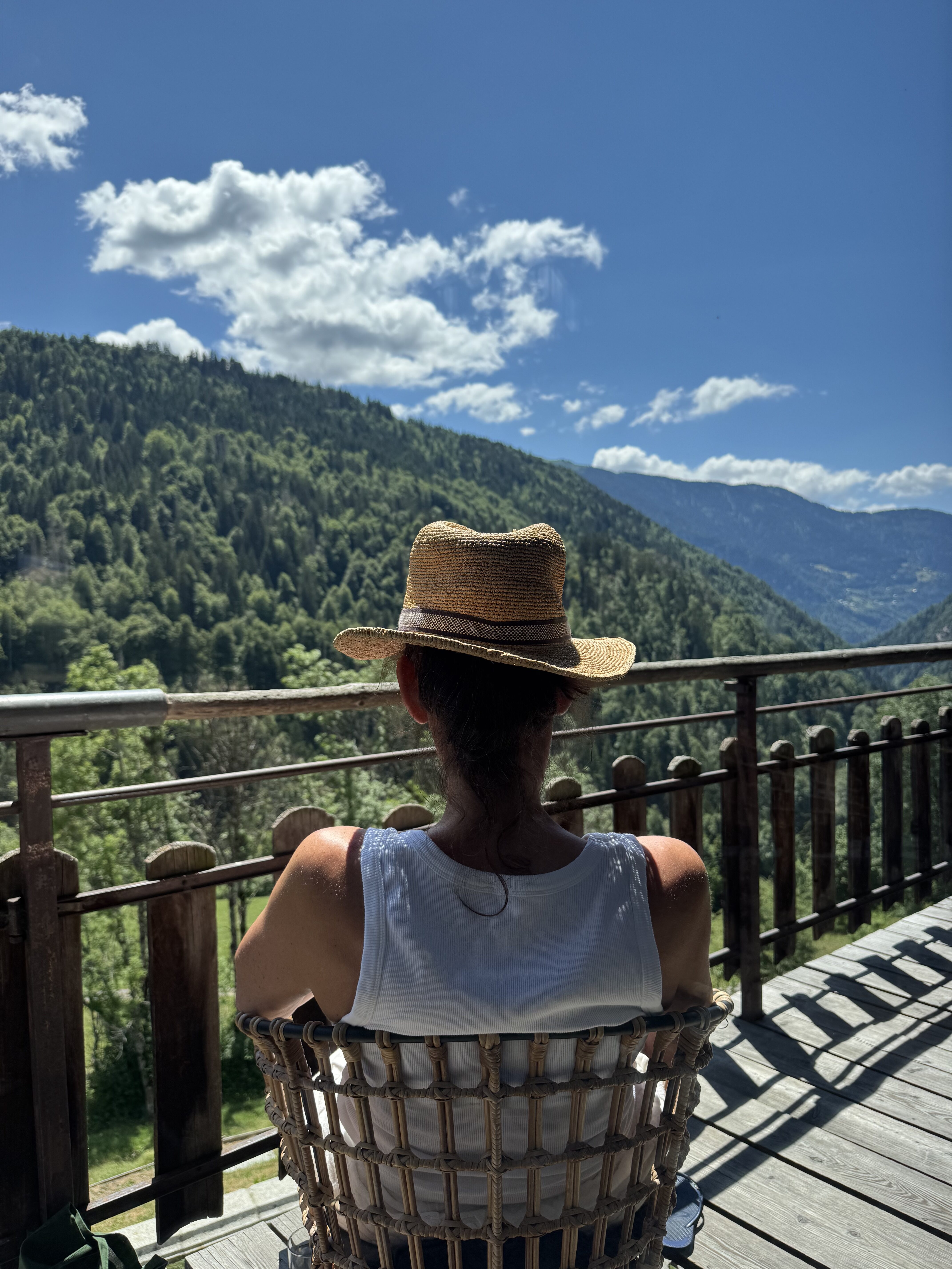 Personne assise sur le balcon en bois du chalet, chapeau de paille, contemplant la vue sur la forêt et les montagnes.