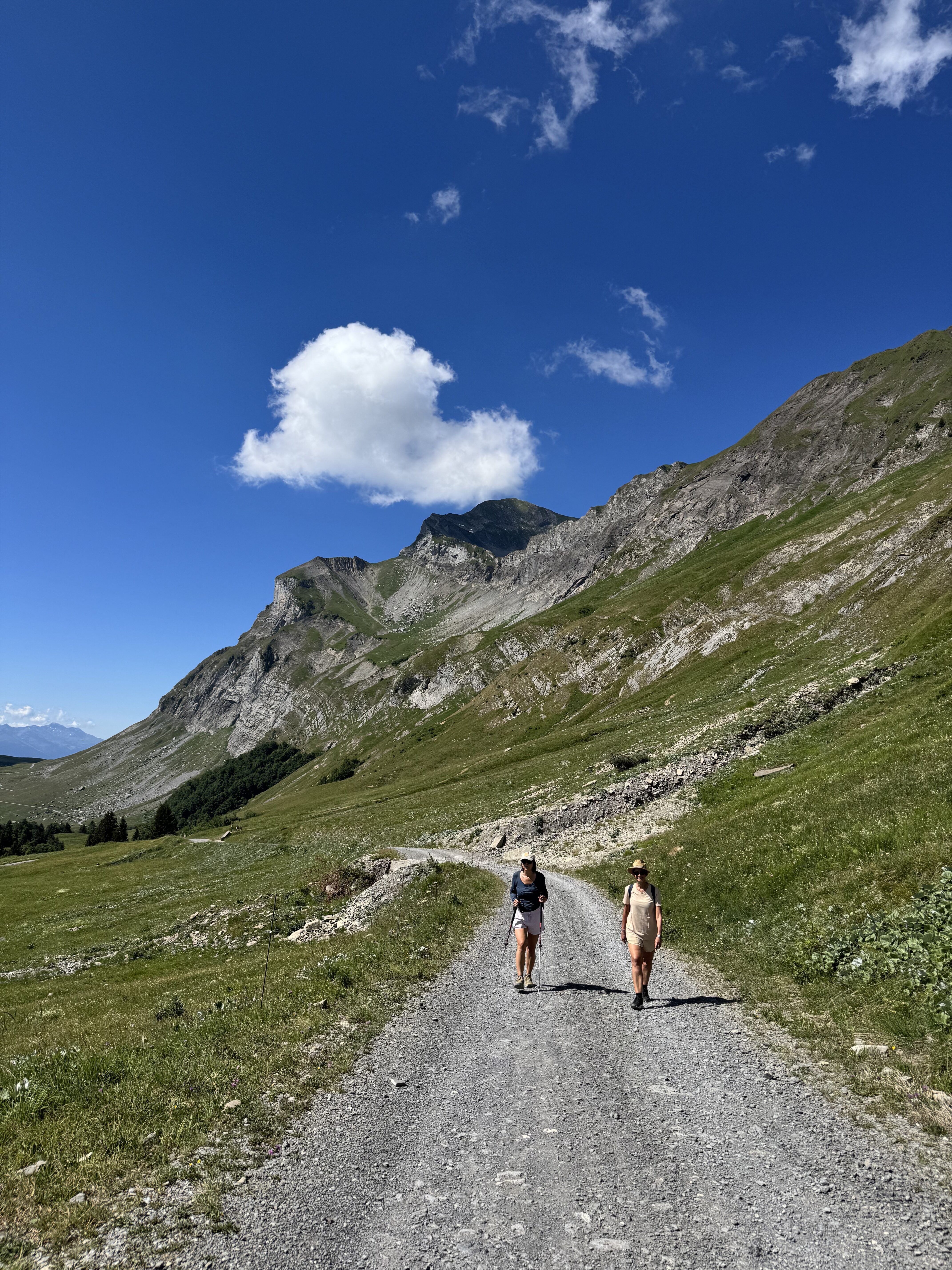 Deux randonneuses marchant sur un chemin de montagne en été, entourées de sommets alpins sous un ciel bleu.