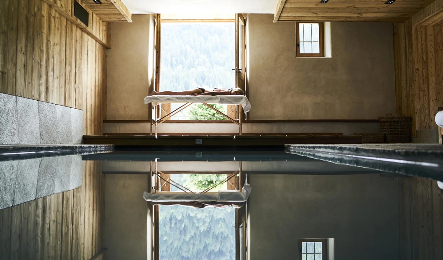 Table de massage installée devant une grande fenêtre donnant sur une forêt de montagne, au bord d'une piscine intérieure au calme miroir. Le reflet de la table et de la fenêtre se dessine parfaitement dans l'eau. Cadre spa alpin haut de gamme en bois et enduit naturel.